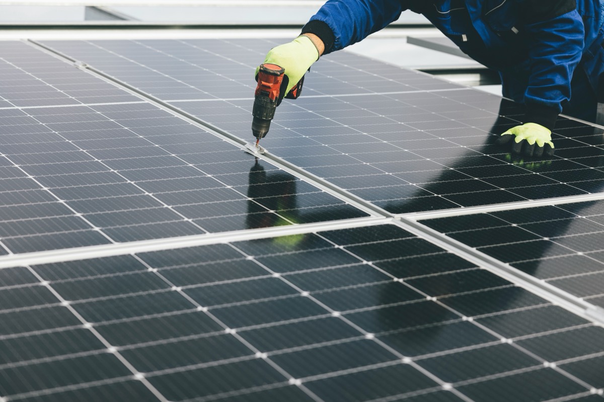 A worker in gloves and blue overalls using a cordless drill to fix a clamp between two solar panels
