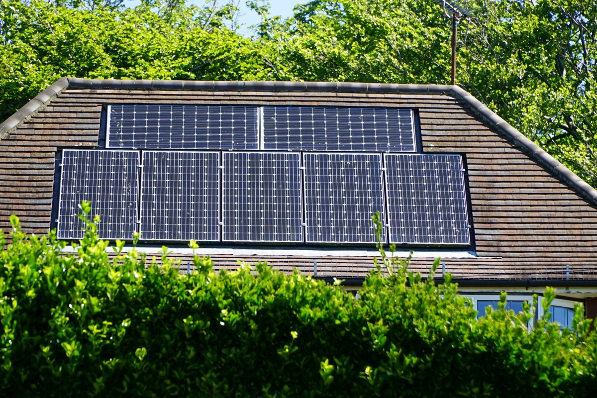 Seven solar panels set into a tiled house roof, with trees behind and a hedge in front